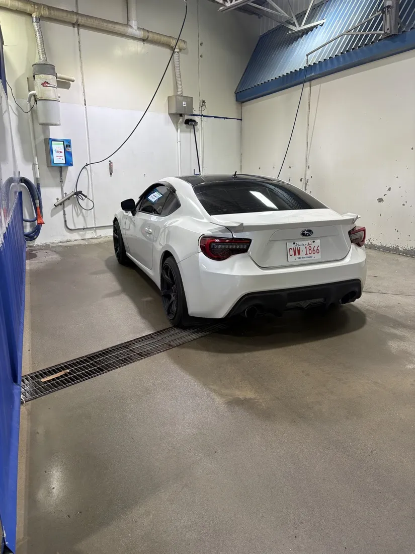 A white car parked in a self-serve bay, with the touchscreen pay terminal, the wand and the vacuum hose on the wall beside it