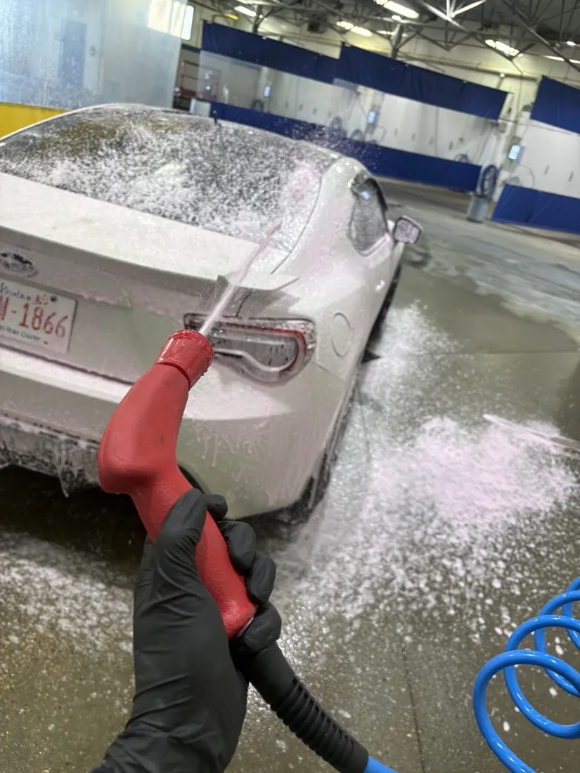 A gloved hand foaming the back of a white car with the red wand, the curtained row of self-serve bays behind it